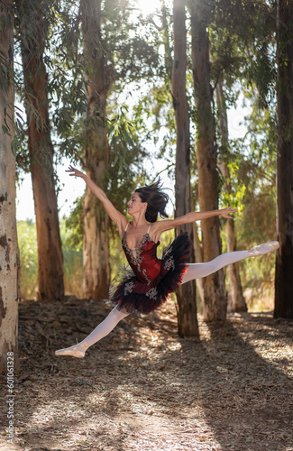 slim classical ballet dancer in a red, white and black dress in a forest with dry leaves on the ground, performing a great acrobatic leap with her legs wide open and stretched