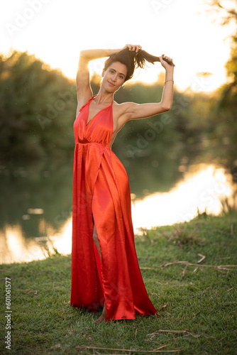 beautiful latin woman holding her hair, wearing an elegant red dress, in a beautiful natural landscape by a lake at sunset with the sunlight reflecting on the water.