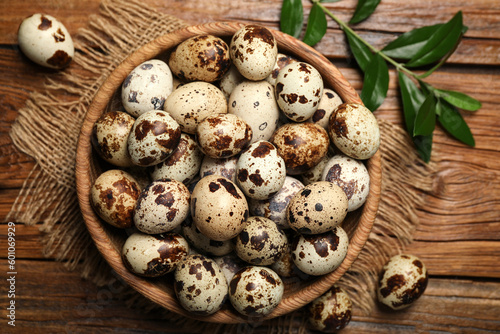 Bowl with quail eggs and green leaves on wooden table, flat lay