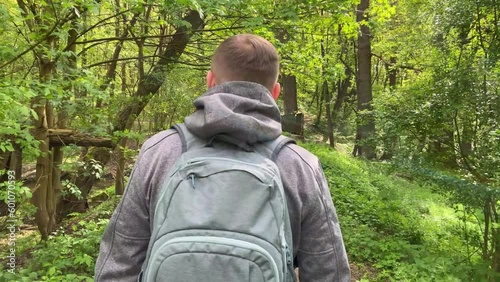 Slow motion camera follows man hiker in grey hooded jacket with backpack. Attractive happy young man with backpack hiking in green wood at sunny spring day.