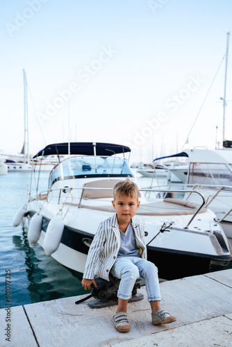 Cute stylish boy sitting on the pier near yacht in marina