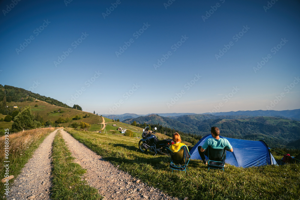 A pair of motorcyclists enjoy the view, man and woman. Loving couple of tourists. Relationship and adventure concept. A touring motorcycle, tent. Active lifestyle. Folding chairs. Copy space