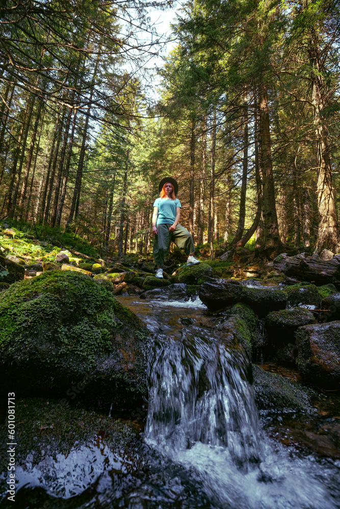 A woman explores new, magical, and fantastic places around the world, surrounded by nature. Female hiker crossing the river. Coniferous forest, tall Carpathian spruces. Vertical photo