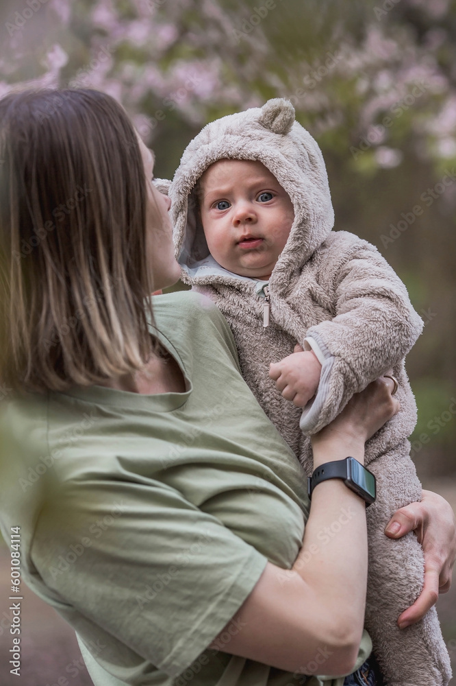 Fototapeta premium A charming boy in nature among flowering trees in his mother's arms. A child of 4 months plays with his mother and smiles. Family, pregnancy and childbirth.