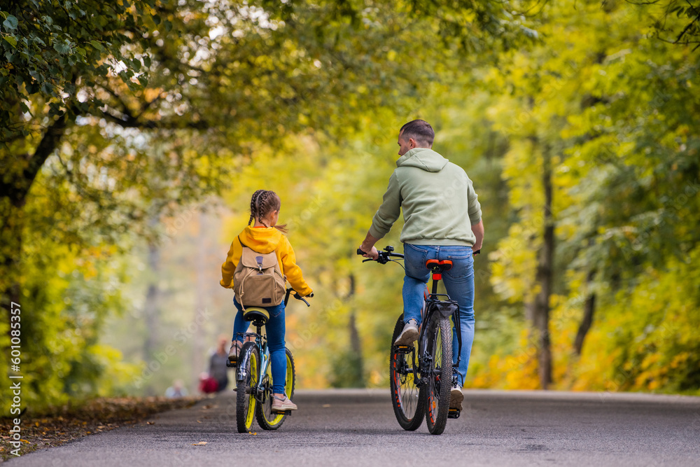 Happy father and daughter ride bicycles in autumn park on sunny day.