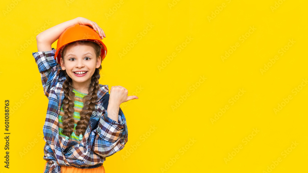 A little girl in a hard hat points to your advertisement on a yellow isolated background. A child in the clothes of a worker, preparing for repairs. Copy space. Banner. A place for advertising. - obrazy, fototapety, plakaty 
