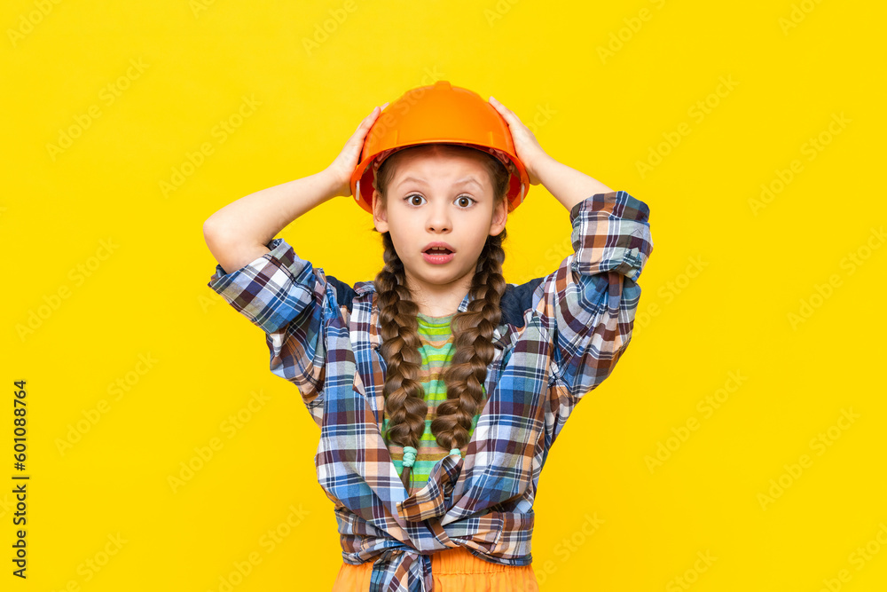 A child in a construction helmet holds his head in surprise. A little girl is getting ready for renovation in the children's room. The profession of an engineer. Yellow isolated background.