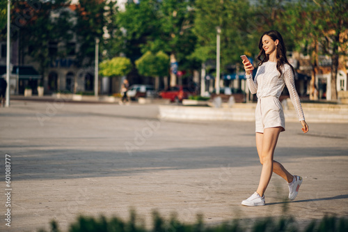 A cheerful young woman in summer fashion style clothes is walking on the city street on a sunny summer day and using her phone.