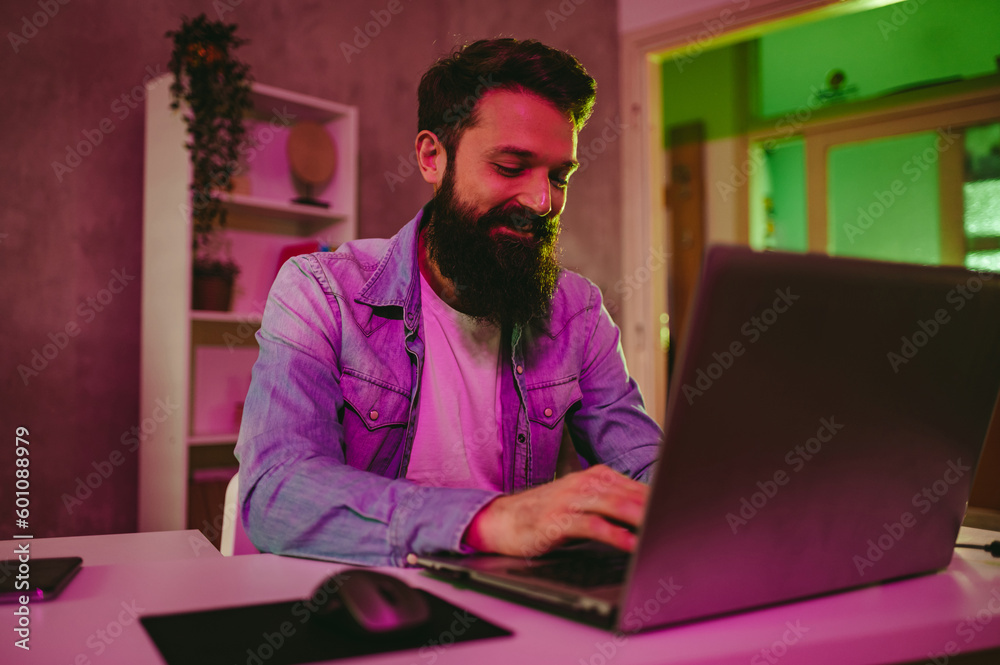 A happy IT specialist is typing on his laptop at night. Neon purple lighting is on. A happy IT specialist is typing on his laptop at night. Neon purple lighting is on.