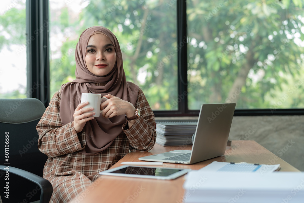 Portrait, Beautiful and attractive millennial Asian Muslim woman wearing hijab enjoying her coffee while working at her office desk. Portrait, Beautiful and attractive millennial Asian Muslim woman wearing hijab enjoying her coffee while working at her office desk.