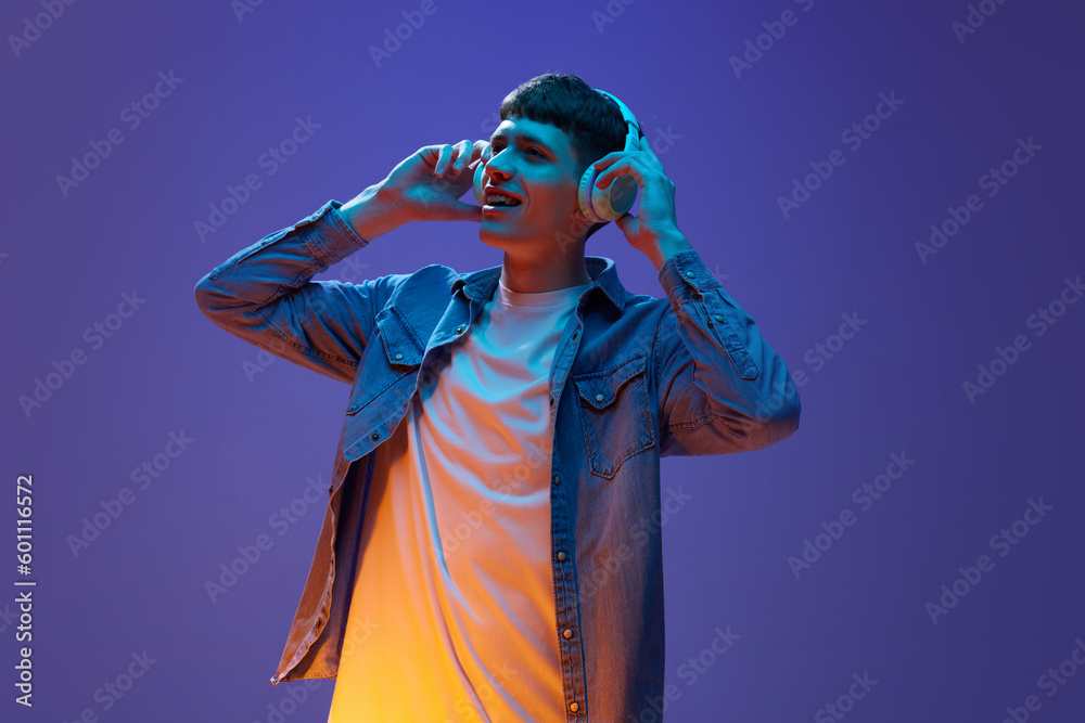 Fototapeta premium Portrait of young relaxed man in casual clothes, listening to music in headphones, having fun against gradient purple background in neon light. Concept of human emotions, lifestyle, youth