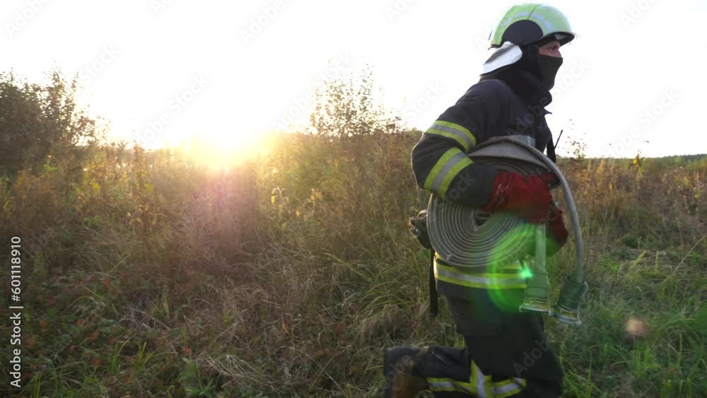 Male firefighter in full equipment running with fire hose to place of emergency. Young fireman in uniform jogging through lawn to extinguishes fire. Concept of environmental issues and global warming Male firefighter in full equipment running with fire hose to place of emergency. Young fireman in uniform jogging through lawn to extinguishes fire. Concept of environmental issues and global warming