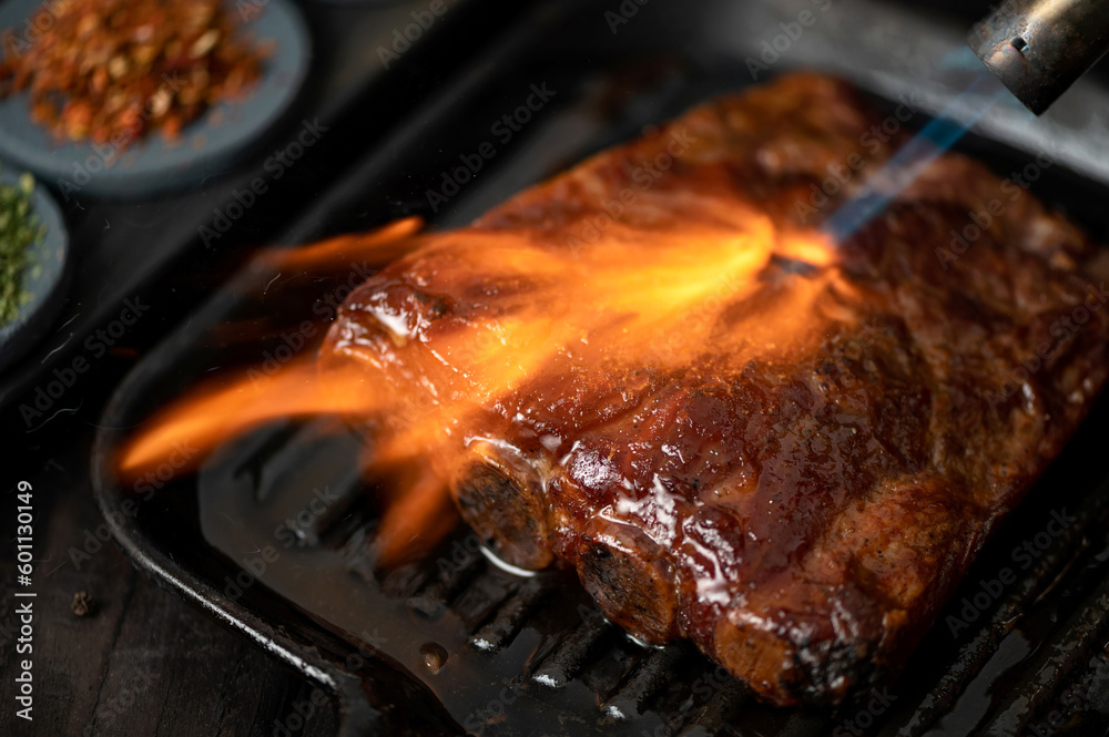 Smoked pork ribs on grill pan under stream of fire. Grilled ribs. Ribs in barbecue sauce. Meat in frying pan on wooden table. Close-up. View from above. Soft focus. Smoked pork ribs on grill pan under stream of fire. Grilled ribs. Ribs in barbecue sauce. Meat in frying pan on wooden table. Close-up. View from above. Soft focus.