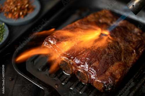 Smoked pork ribs on grill pan under stream of fire. Grilled ribs. Ribs in barbecue sauce. Meat in frying pan on wooden table. Close-up. View from above. Soft focus.