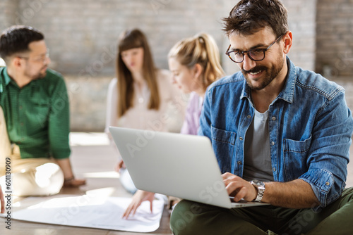 Young entrepreneur using computer while sitting on the floor in the office. There are people in the background