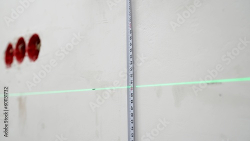 A man uses a cross laser level and a tape for accurate measurements on a light wall, close-up. Measure the height with a laser level. Laser level and tape measure in action. © Ruslan