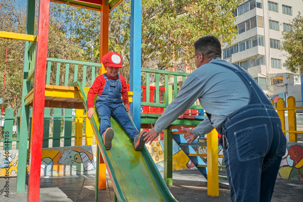 latino boy playing on the slide with his dad dressed as a video game in an outdoor park in the city of la paz bolivia.
