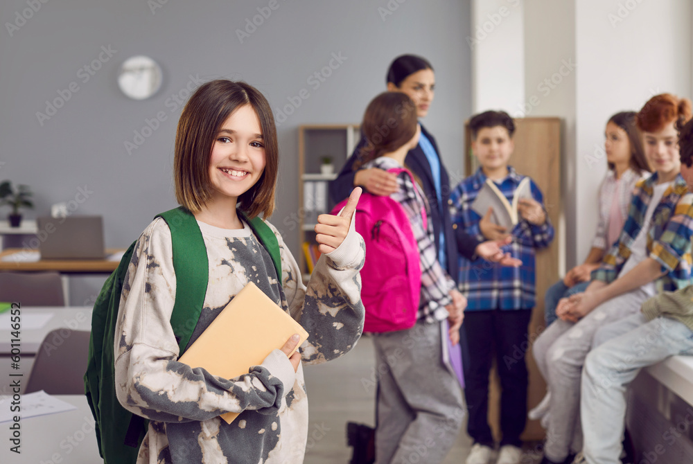 Fototapeta premium Portrait of happy girl school student showing thumb up. Beautiful smiling schoolgirl wearing casual clothes standing with backpack on background of her classmates in classroom and looking at camera