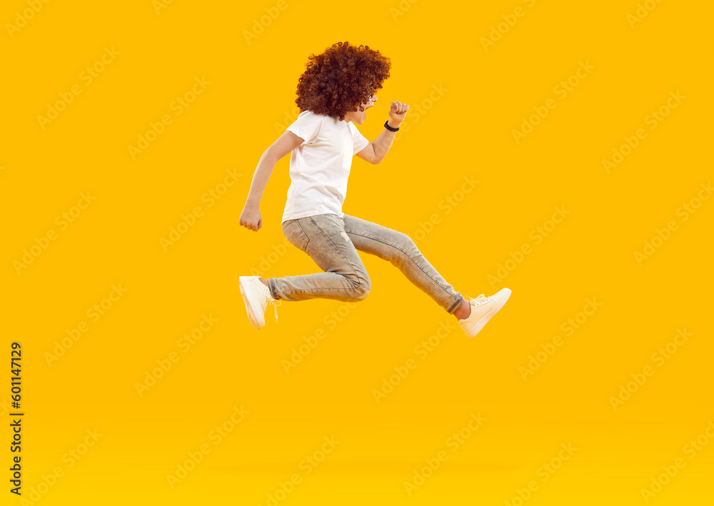 Energetic, funny, happy boy in red - haired curly wig jumps and screams with his mouth open, dressed in white T-shirt and gray jeans, on an isolated yellow-orange background. Run and jump.