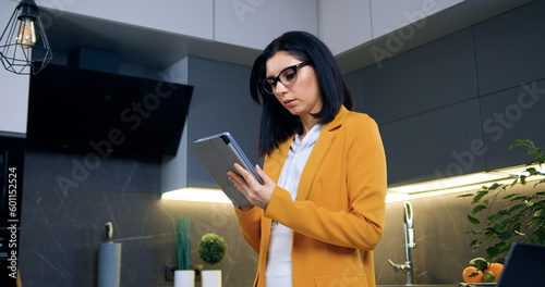 Attractive serious busy 35-aged brunette in eyeglasses dressed in trendy clothes working on tablet pc standing in beautifully decorated cuisine,front view