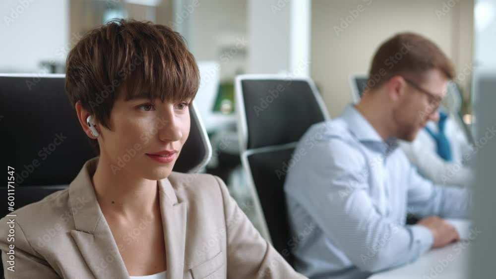 Pretty Caucasian woman manager with suit using earpods in office. Young busy female sitting in front of computer while listening music. In blurred background men working on PC. Technology concept. Pretty Caucasian woman manager with suit using earpods in office. Young busy female sitting in front of computer while listening music. In blurred background men working on PC. Technology concept.