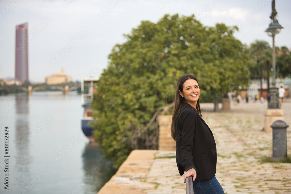 Young beautiful woman with straight brown hair, jacket and jeans leaning on a railing by the river, smiling happily. Concept fashion, beauty, trend, millennial, travel.