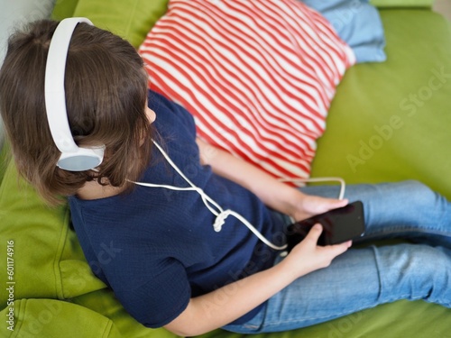 little girl is lying on a sofa in headphones and looking into a mobile phone