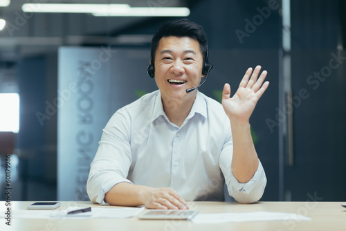 Portrait of a smiling Asian businessman talking on a video call in a headset, greeting the camera, sitting in the office at the table. Gestures, advises, explains, helps.