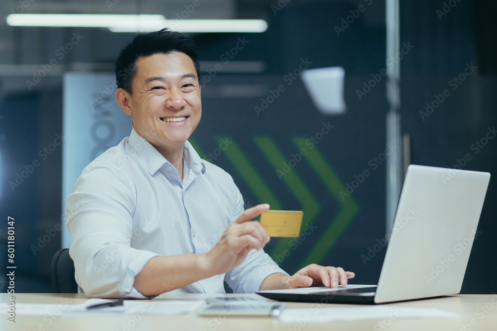 Financial transactions with a credit card. Young Asian man working in office at laptop and using credit card, posing and smiling at camera. Financial transactions with a credit card. Young Asian man working in office at laptop and using credit card, posing and smiling at camera.