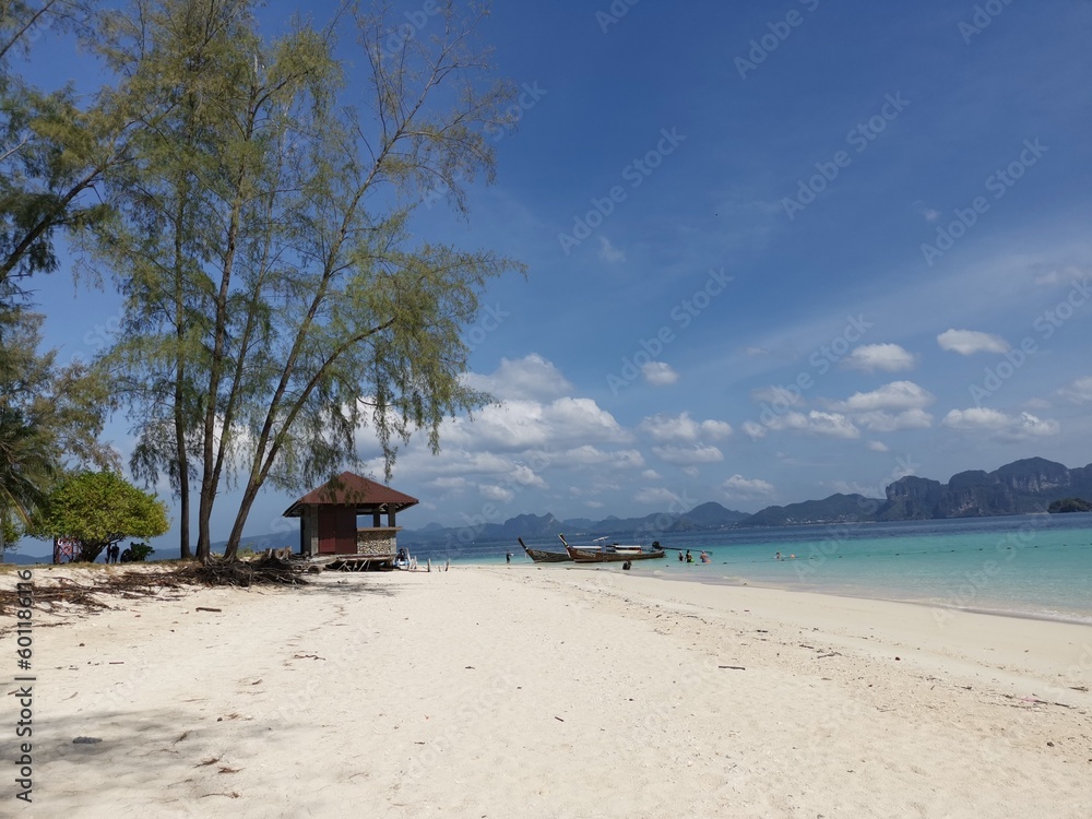 Beach rocks. Two boats on the shore near the pavilion for rest. Trees provide shady and shade. Green sea, white sand, blue sky with light clouds. Islands and mountains in background.