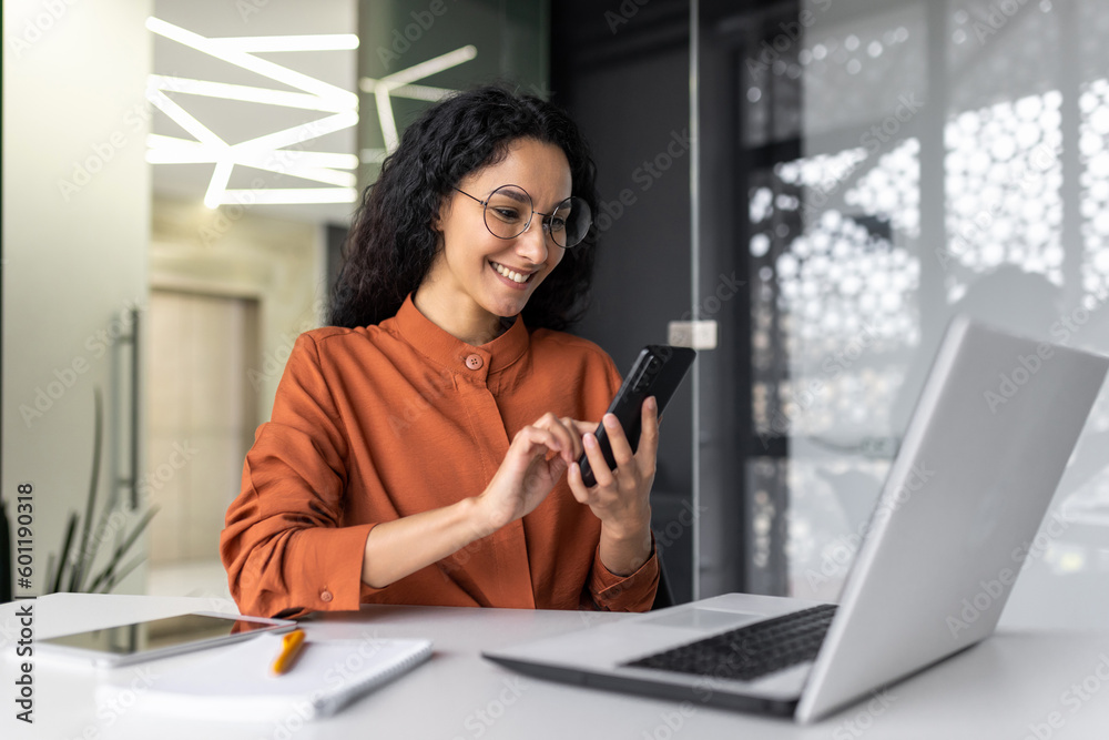 Beautiful woman in the office, happy and smiling latin american business woman uses internet phone close up, female worker reads message and browses internet pages inside office building - obrazy, fototapety, plakaty 