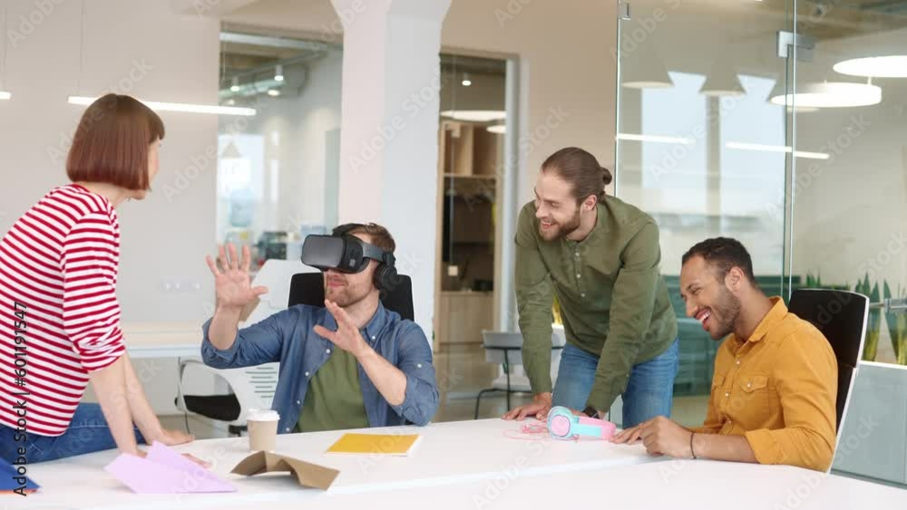 Group of mixed-raced people sitting in office and communicating with each other. Funny man wearing virtual reality headset and checking look of future office. Workers smiling and laughing together. Group of mixed-raced people sitting in office and communicating with each other. Funny man wearing virtual reality headset and checking look of future office. Workers smiling and laughing together.