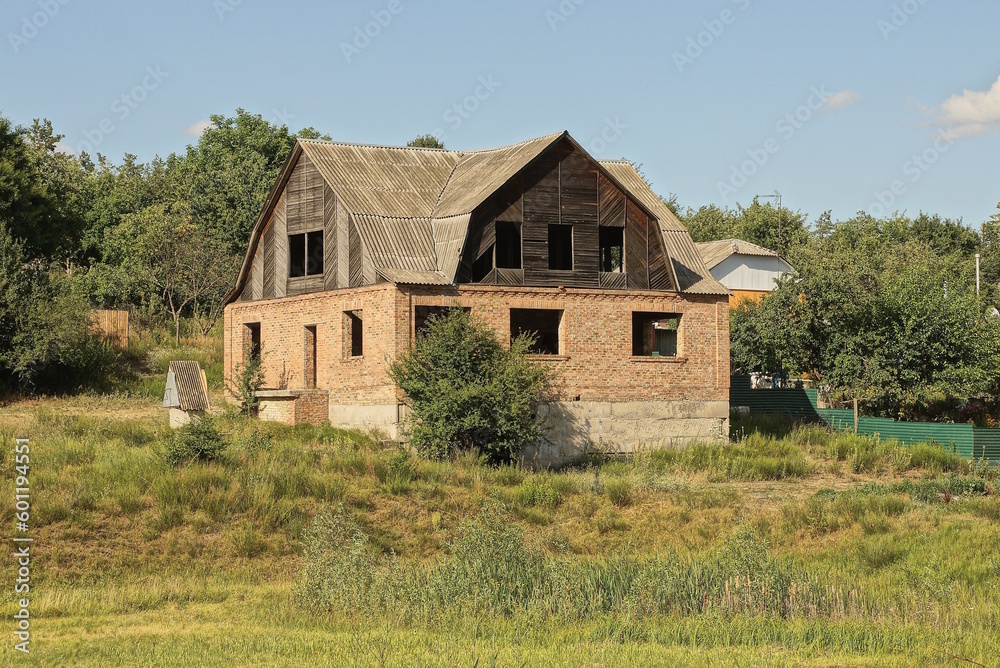 one brown unfinished brick house with empty windows and a black wooden attic on the street among green grass and vegetation one brown unfinished brick house with empty windows and a black wooden attic on the street among green grass and vegetation