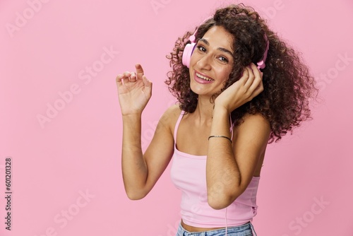 Happy woman wearing headphones with curly hair listening to music and singing along with her eyes closed in a pink T-shirt and jeans on a pink background DJ party, copy space