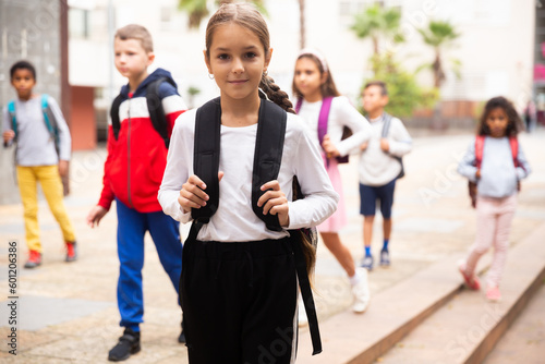 Portrait of positive schoolgirl standing near school, children on background