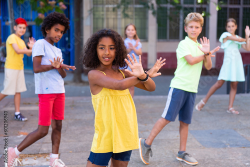 Smiling curly african american preteen girl dancing modern choreography with group of tweenagers on city street on summer day..