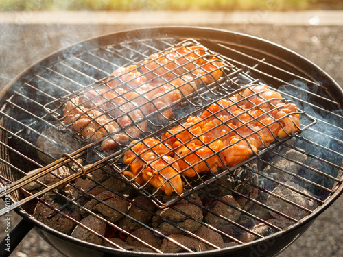 Cooking marinated chicken in a metal basket on small round grill. Sumer time activity, Preparing food on fire in a garden or park. Barbeque time.