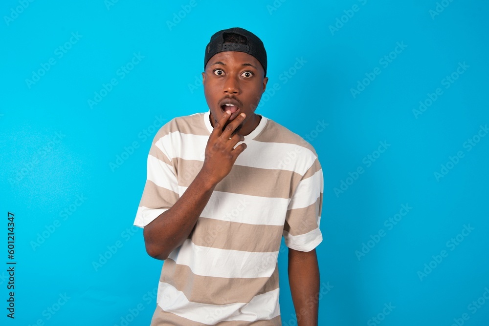 Nervous puzzled Handsome man wearing striped t-shirt and cap over blue background opens mouth from surprise, reacts on sudden news.