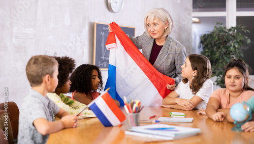 Friendly high school teacher, conducting a lesson in the classroom, tells pupils the history of the Netherlands and holds the .national flag of the country in her hands