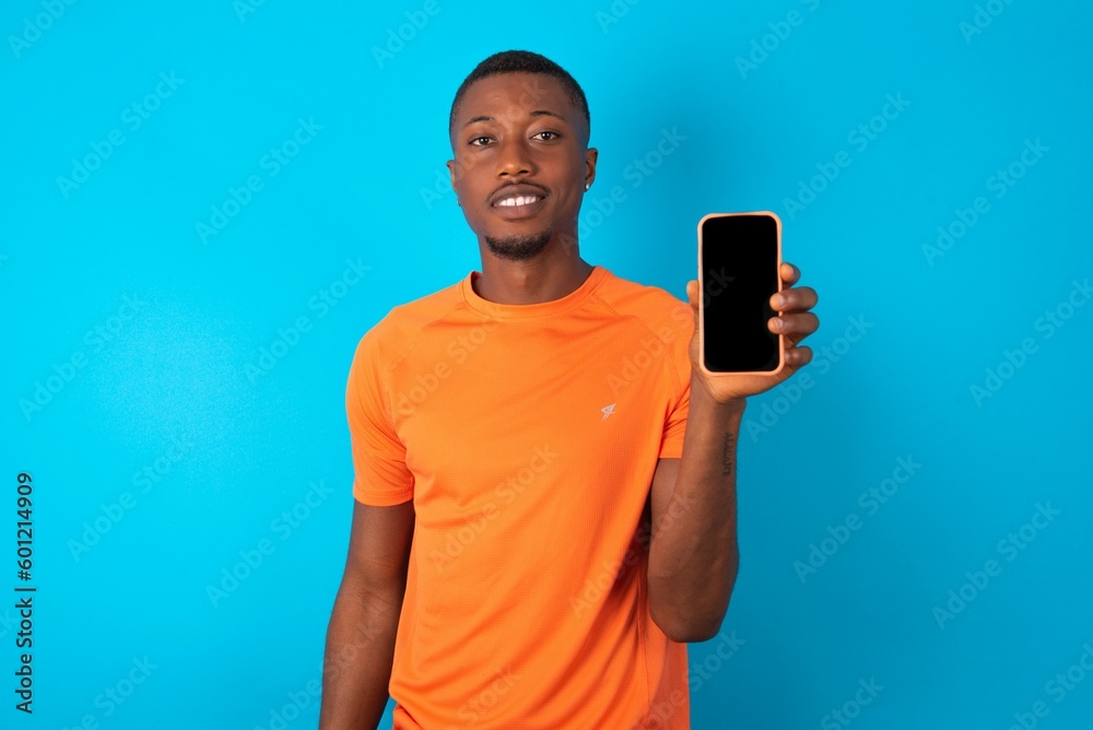 Smiling Handsome man wearing orange T-shirt over blue background Mock up copy space. Hold mobile phone with blank empty screen Smiling Handsome man wearing orange T-shirt over blue background Mock up copy space. Hold mobile phone with blank empty screen