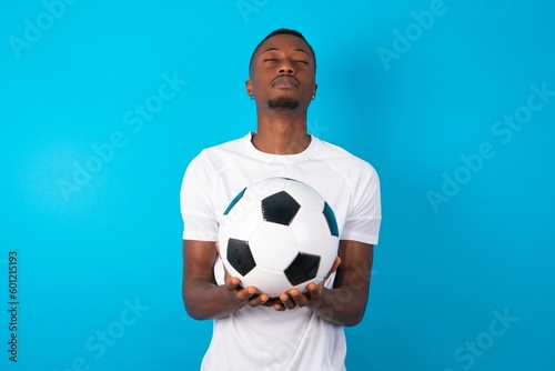 Man wearing white T-shirt holding a ball over blue background nice-looking sweet charming cute attractive lovely winsome sweet peaceful closed eyes