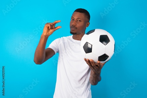 Displeased Man wearing white T-shirt holding a ball over blue background shapes little hand sign demonstrates something not very big. Body language concept.