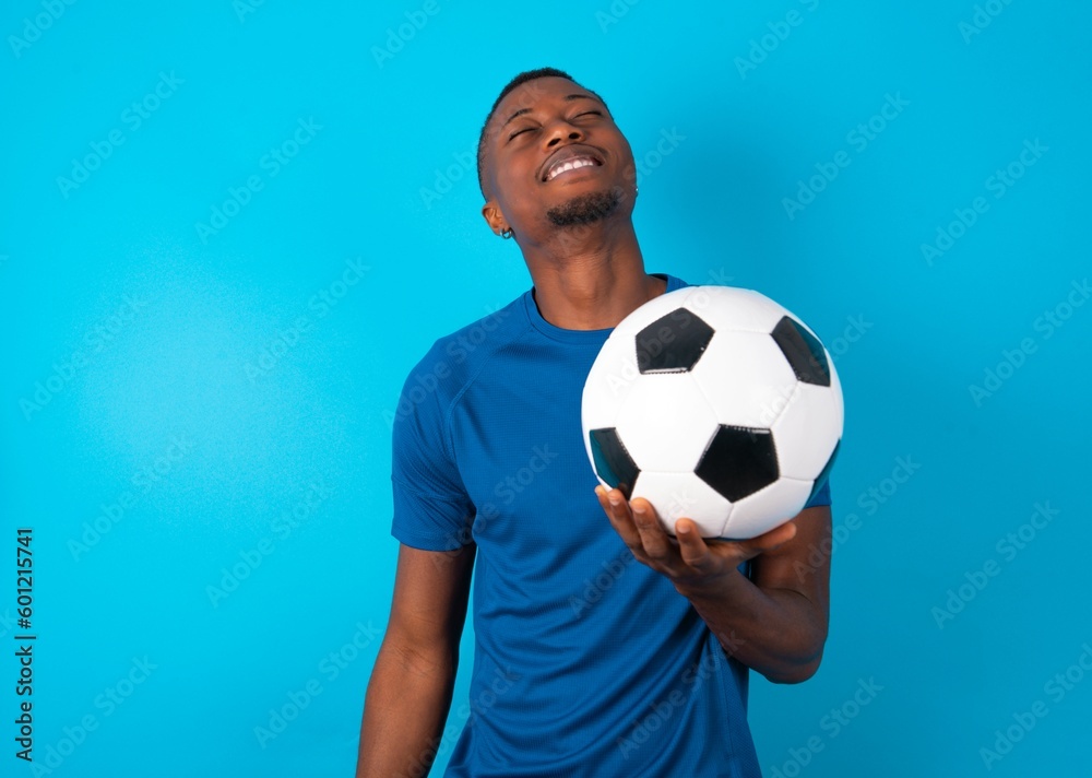 Positive Young man wearing blue T-shirt holding a ball over blue background with overjoyed expression closes eyes and laughs shows white perfect teeth Positive Young man wearing blue T-shirt holding a ball over blue background with overjoyed expression closes eyes and laughs shows white perfect teeth