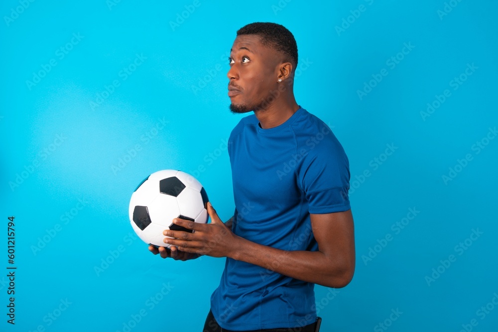 Young man wearing sport T-shirt holding a ball over blue background stares aside with wondered expression has speechless expression. Embarrassed model looks in surprise Young man wearing sport T-shirt holding a ball over blue background stares aside with wondered expression has speechless expression. Embarrassed model looks in surprise