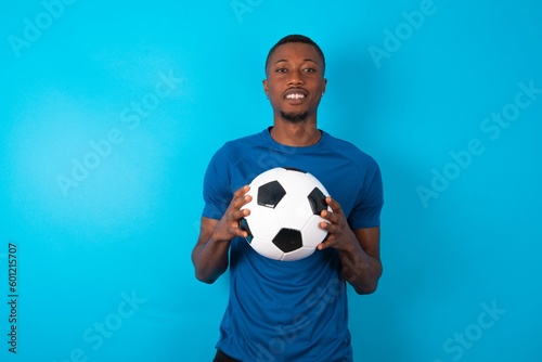 Young man wearing blue T-shirt holding a ball over blue background with nice beaming smile pleased expression. Positive emotions concept