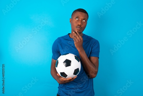 Thoughtful Young man wearing blue T-shirt holding a ball over blue background holds chin and looks away pensively makes up great plan