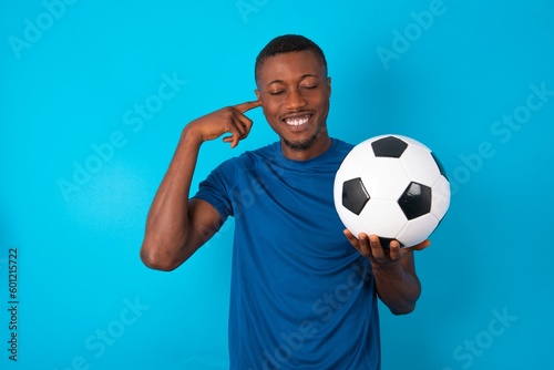 Happy Young man wearing blue T-shirt holding a ball over blue background ignores loud music and plugs ears with fingers asks to turn off sound