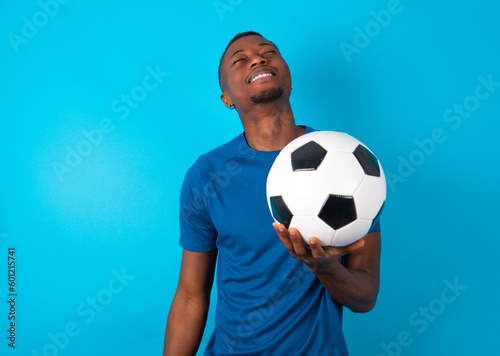 Positive Young man wearing blue T-shirt holding a ball over blue background with overjoyed expression closes eyes and laughs shows white perfect teeth © Roquillo