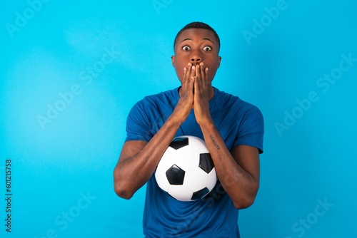 Young man wearing sport T-shirt holding a ball over blue background keeps hands on mouth, looks with eyes full of disbelief, being puzzled with amount of work