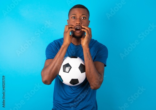 Speechless Young man wearing sport T-shirt holding a ball over blue background keeps hands near opened mouth reacts to shocking news stares wondered at camera